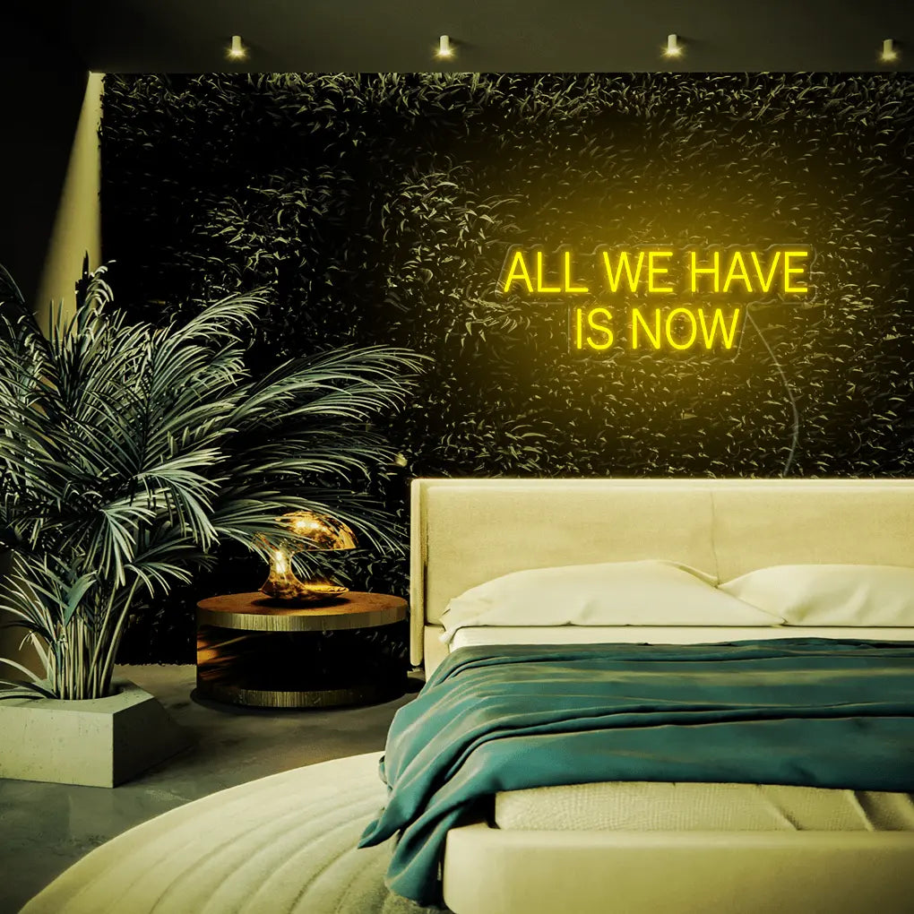 All We Have Is Now LED Neon Sign in yellow, featuring block lettering above a bed beside indoor plants.