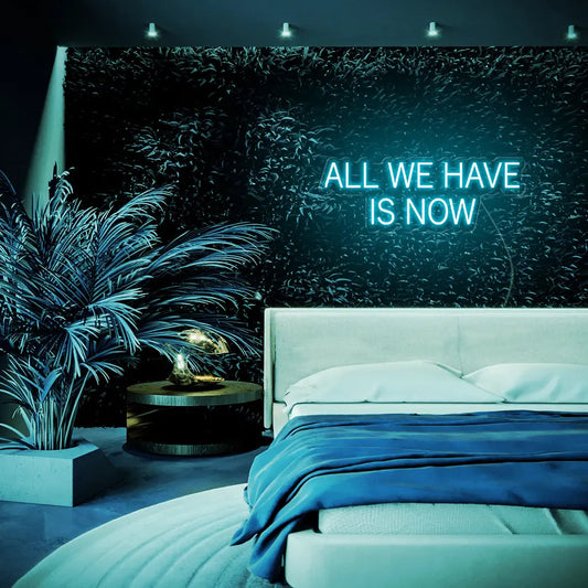 All We Have Is Now LED Neon Sign in turquoise, featuring block lettering above a bed beside indoor plants.