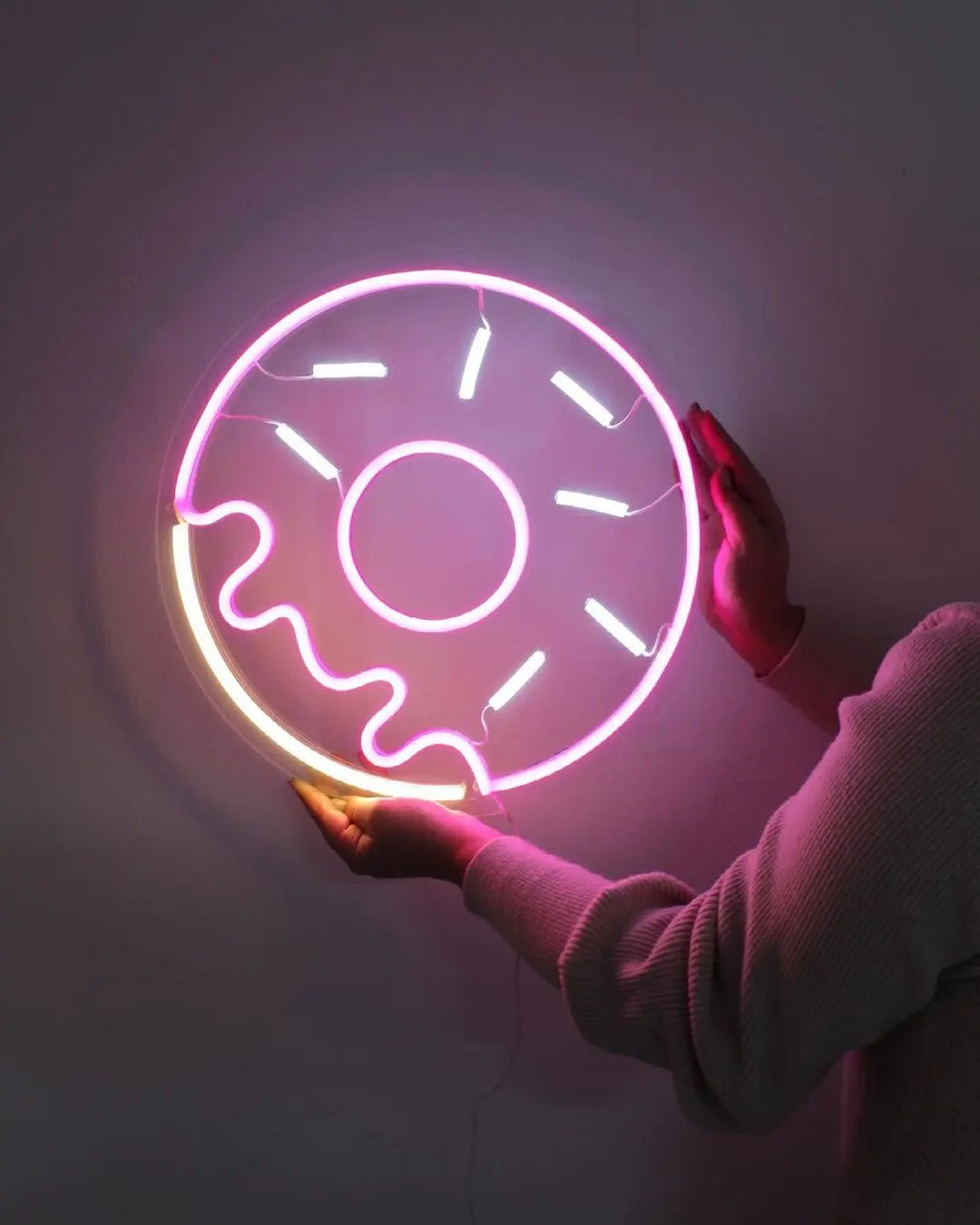 Donut LED Neon Light Sign in pink and white, featuring outlined iced doughnut held against a dark wall.