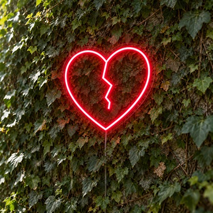 Broken Heart LED Neon Light Sign in red, featuring broken-heart outline against a green foliage wall.
