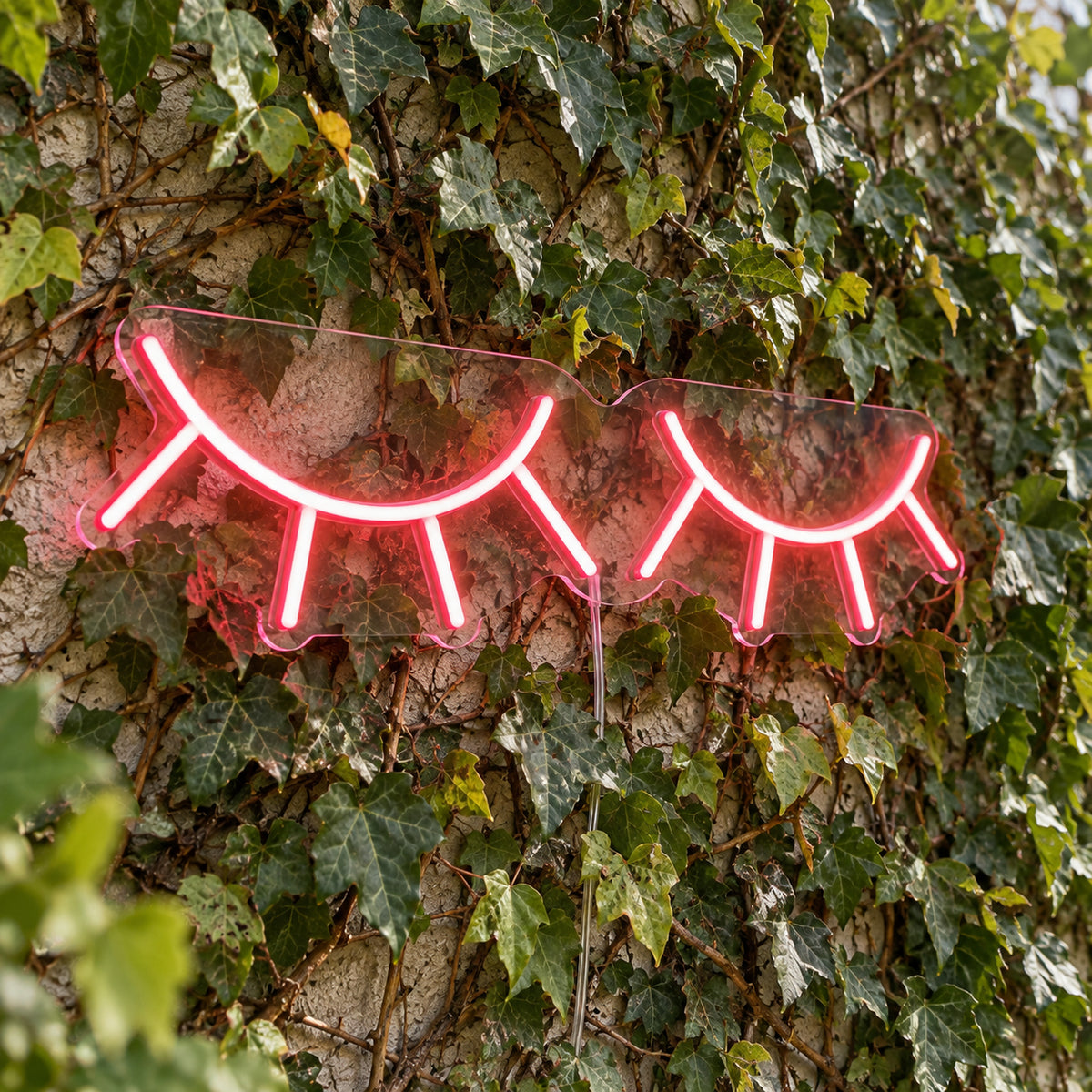 Eyelashes LED Neon Light Sign in pink, featuring two closed eyelash outlines against a green foliage wall.