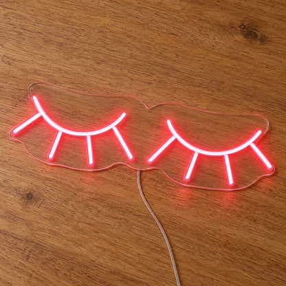Eyelashes LED Neon Light Sign in pink, featuring two closed eyelash outlines on a wooden surface.