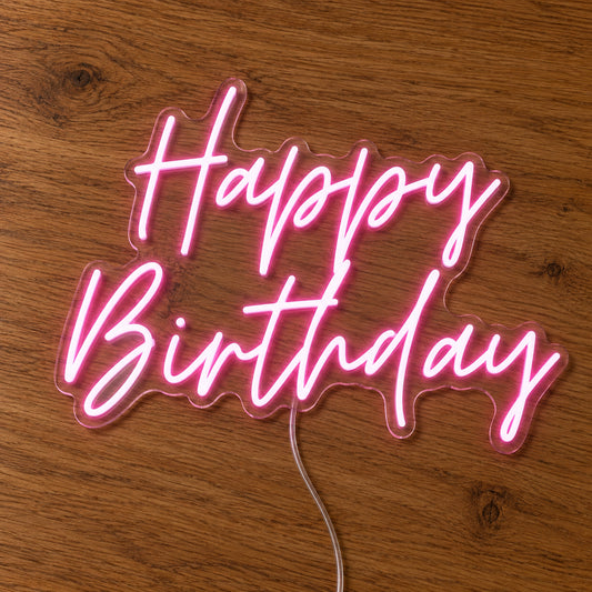 Happy Birthday LED Neon Light Sign in pink, featuring two lines of script lettering on a wooden surface.