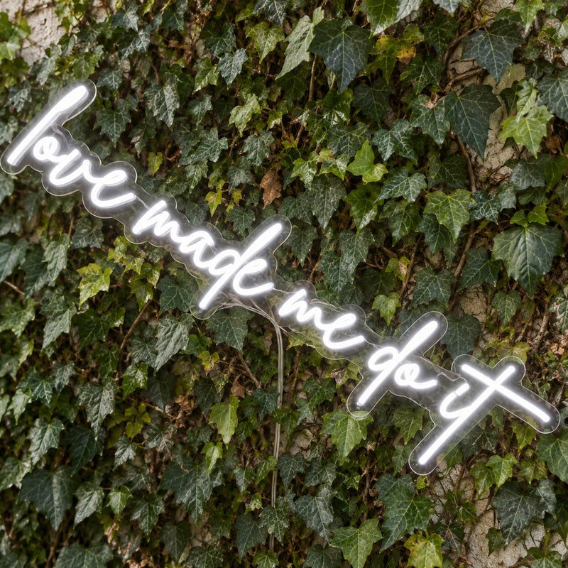 Love Made Me Do It LED Neon Light Sign in white, featuring script lettering against a green foliage wall, viewed at an angle.
