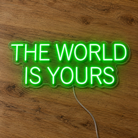 The World Is Yours Neon Sign in green, featuring two lines of block lettering on a wooden surface.