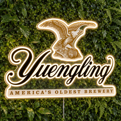 Yuengling LED Neon Sign, featuring illuminated beer-brand logo on a green foliage wall.