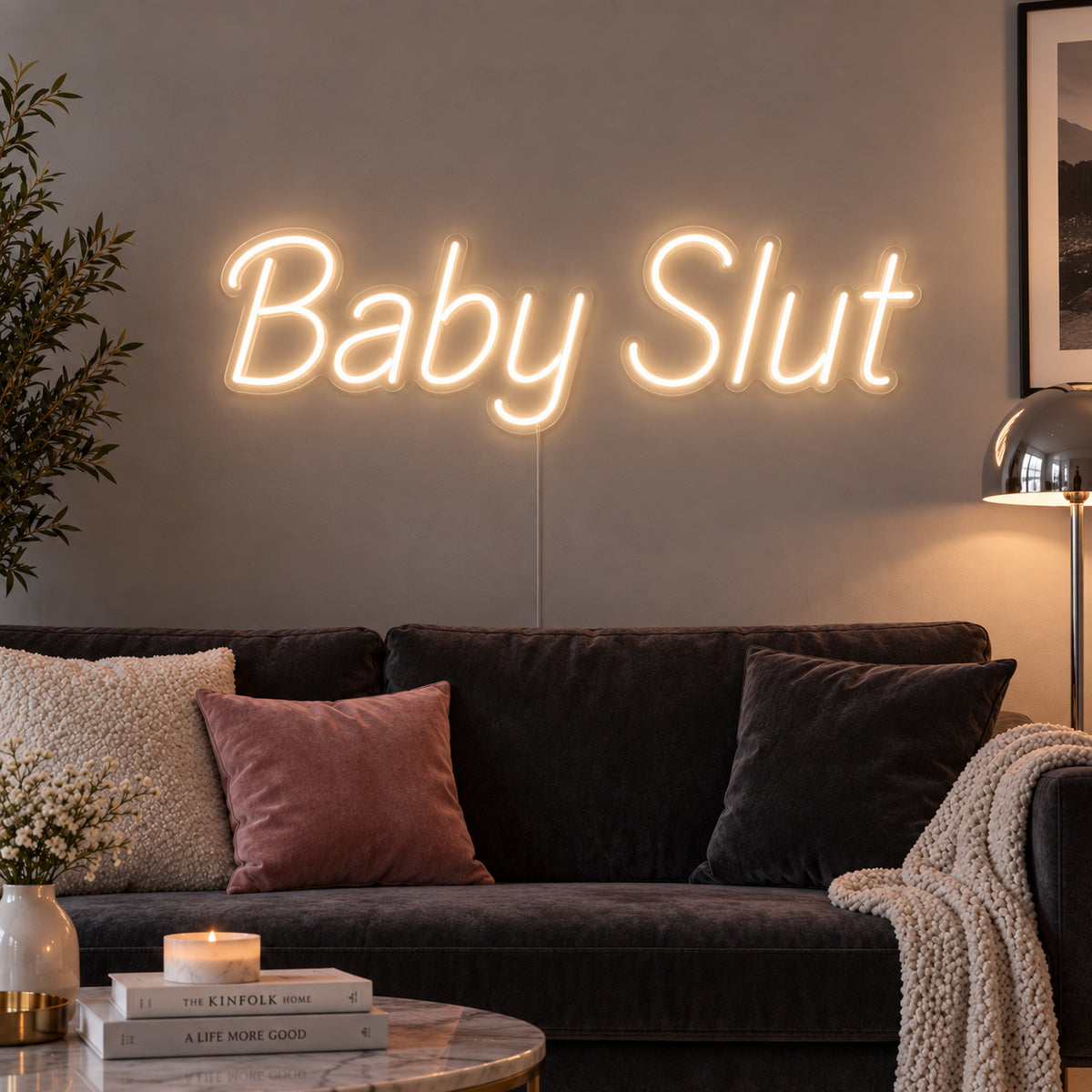 Warm White version of the Baby Slut Neon Sign, shown above a grey sofa with pink cushions.