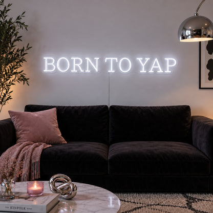 Born To Yap Neon Sign glowing white above a dark sofa and glass coffee table.