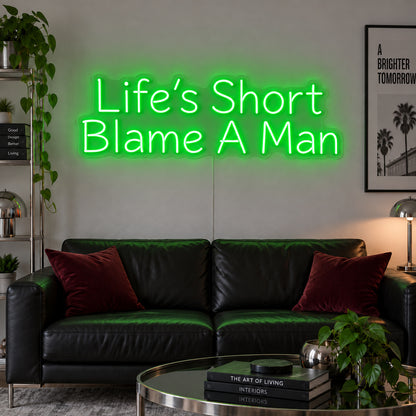 Life's Short Blame A Man Neon Sign with green lighting, displayed above a black leather sofa.