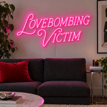 Lovebombing Victim Neon Sign glowing pink above a dark sofa with red cushions.