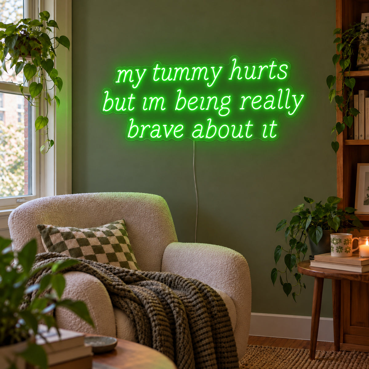 My Tummy Hurts But Im Being Really Brave About It Neon Sign with green lighting, displayed above a reading chair against a green wall.