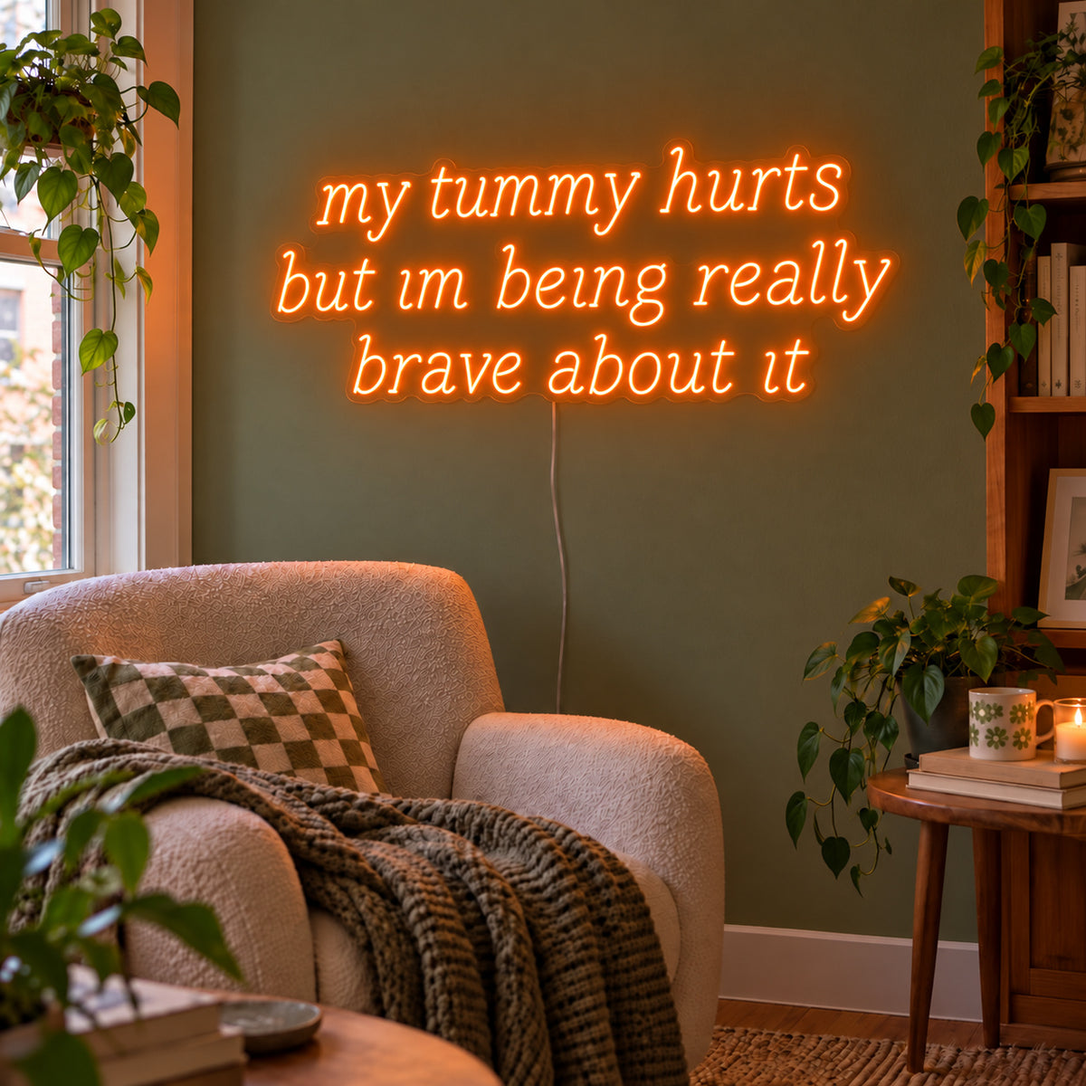 Orange version of the My Tummy Hurts But Im Being Really Brave About It Neon Sign, shown above a reading chair against a green wall.