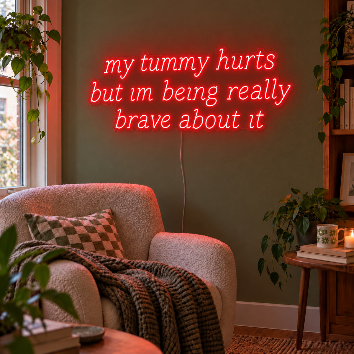 My Tummy Hurts But Im Being Really Brave About It Neon Sign with red lighting, displayed above a reading chair against a green wall.