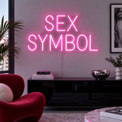 Sex Symbol Neon Sign in pink, featuring two lines of Sex Symbol block lettering above a black sideboard.