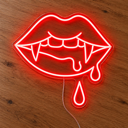 Vampire Lips with Double Blood Drops Neon Sign featuring red lips with pointed fangs and two hanging blood drops, illuminated on clear acrylic lying on a natural oak wooden surface.